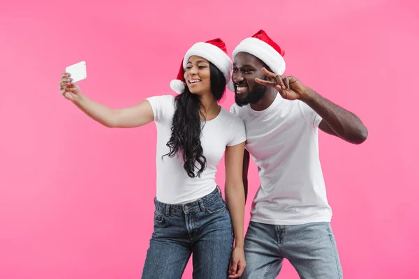 Feliz afroamericana mujer en navidad sombrero tomando selfie en smartphone con novio haciendo paz signo aislado en rosa fondo - foto de stock