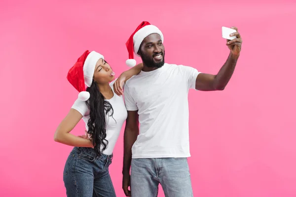Joven afroamericano hombre en navidad sombrero tomando selfie en smartphone con novia pegando lengua fuera aislado en rosa fondo - foto de stock