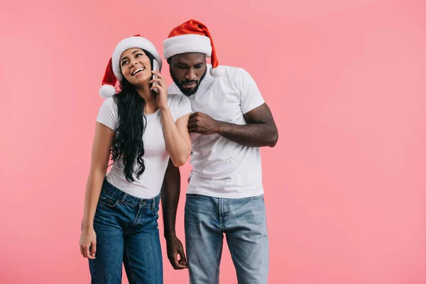 Concentré homme afro-américain dans chapeau de Noël écoutant petite amie parler sur smartphone isolé sur fond rose — Photo de stock