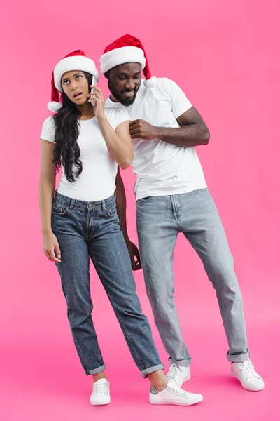 Smiling african american man in chrismas hat eavesdropping girlfriend talking on smartphone on pink background — Stock Photo