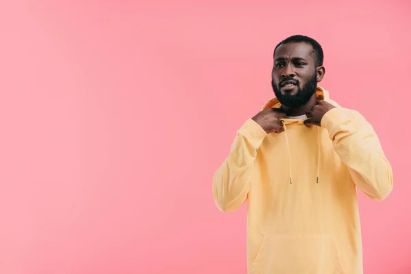 Elegante joven afroamericano hombre quitando sudadera con capucha amarilla aislado sobre fondo rosa - foto de stock