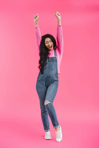 Excited stylish young african american woman in denim overall with raised arms on pink background — Stock Photo