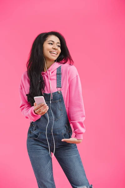 Smiling young african american woman in earphones listening music with smartphone isolated on pink background — Stock Photo