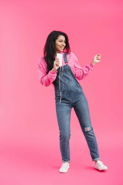 Sonriente mujer afroamericana en auriculares escuchando música con smartphone bailando sobre fondo rosa - foto de stock