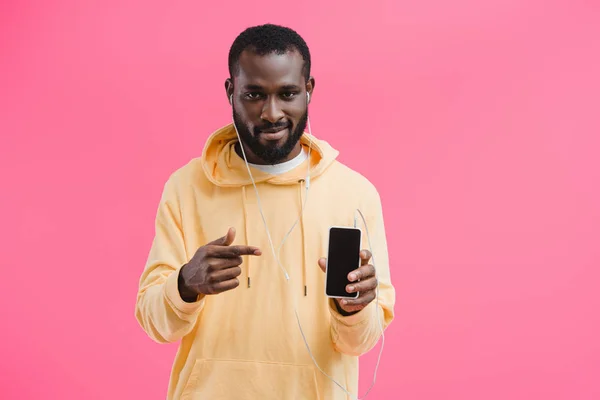 Молодой африканский американец в наушниках, указывающий на смартфон с пустым экраном, изолированным на розовом фоне — стоковое фото