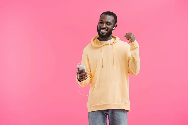 Hombre afroamericano feliz en auriculares gesticulando a mano escuchando música con teléfono inteligente aislado sobre fondo rosa - foto de stock