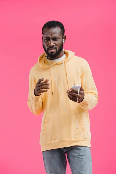 Disappointed african american man in earphones listening music with smartphone isolated on pink background — Stock Photo