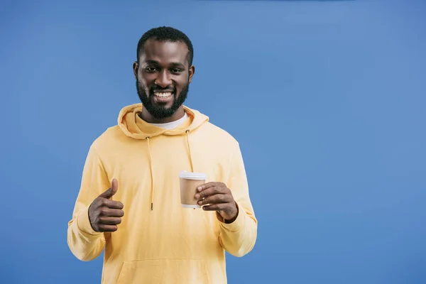 Щасливий молодий афроамериканський чоловік тримає одноразову чашку кави і робить великий палець вгору жест ізольований на синьому фоні — стокове фото
