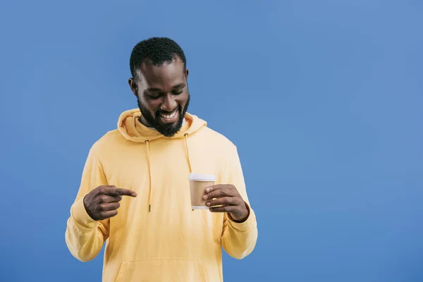 Sorridente jovem afro-americano apontando por dedo para copo de café descartável isolado no fundo azul — Fotografia de Stock