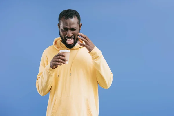 Sconvolto giovane uomo africano americano con smorfia sul viso tenendo monouso tazza di caffè isolato su sfondo blu — Foto stock