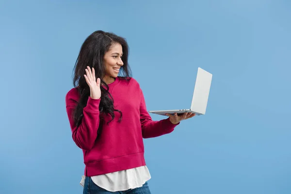 Вид сбоку на счастливую африканскую женщину, ждущую руку и имеющую видеозвонок на ноутбуке, изолированный на синем фоне — стоковое фото