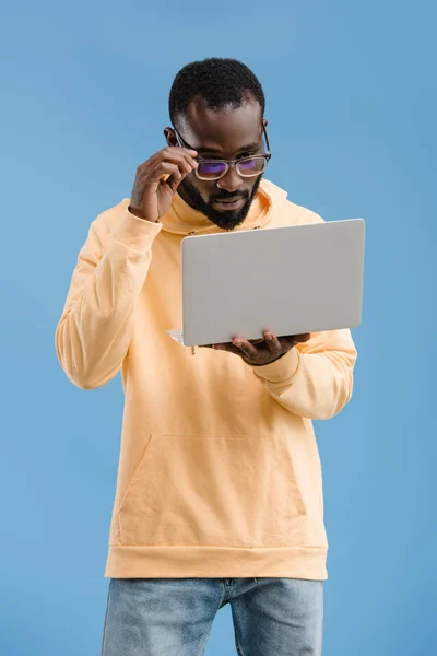 Молодий афроамериканський чоловік регулює окуляри і дивиться на ноутбук ізольовано на синьому фоні — стокове фото