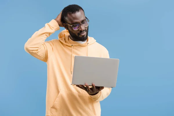 Reflexivo joven afroamericano hombre en gafas mirando portátil aislado sobre fondo azul - foto de stock