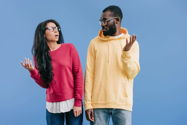 Confuso coppia afroamericana in occhiali da vista guardarsi l'un l'altro e facendo gesto dubio isolato su sfondo blu — Foto stock