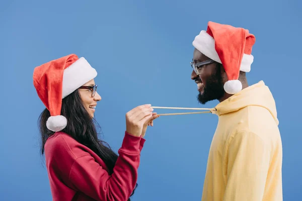 Vista lateral da mulher afro-americana em chapéu de natal segurando atacadores de namorado com capuz isolado em fundo rosa — Fotografia de Stock