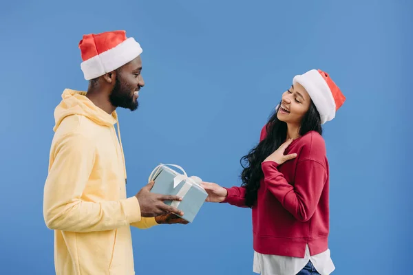 Улыбающийся африканский американец в рождественской шляпе, дарящий подарочную коробку девушке, изолированной на синем фоне — стоковое фото