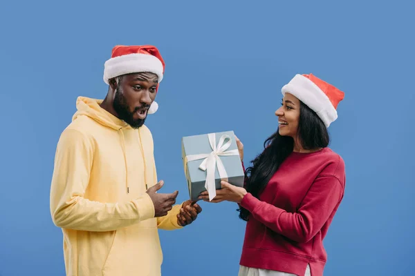Приваблива афроамериканська жінка в різдвяному капелюсі дарує подарункову коробку шокованому хлопцеві ізольовано на синьому фоні — стокове фото
