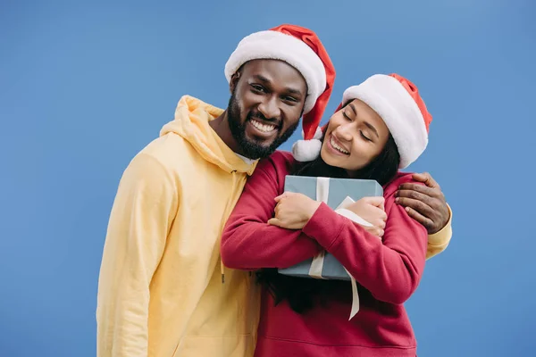 Hombre afroamericano feliz en sombrero de Navidad abrazando novia con caja de regalo aislado sobre fondo azul - foto de stock
