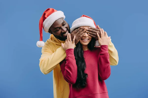 Smiling african american man in christmas hat closing eyes of girlfriend isolated on blue background — Stock Photo