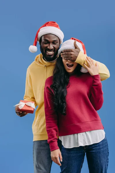 Happy african american man in christmas hat holding gift box and closing eyes of girlfriend isolated on blue background — Stock Photo