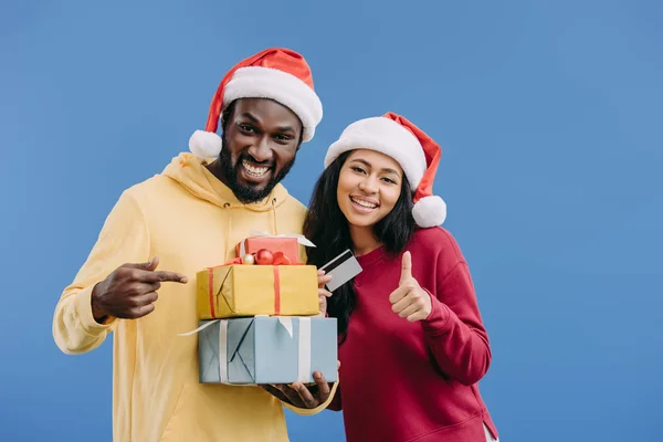 Афроамериканців людини в капелюсі Різдва, вказуючи на подарункові коробки під час його подруга проведення кредитної картки і робить великий палець вгору жестом ізольовані на синьому фоні — стокове фото