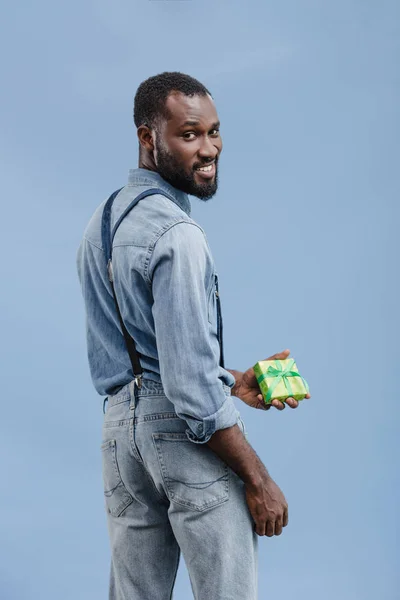 Giovane uomo africano americano con scatola regalo avvolto da nastro guardando la fotocamera isolata su sfondo blu — Foto stock