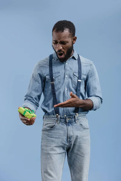 Scioccato giovane uomo africano americano che punta a mano a scatola regalo avvolto da nastro isolato su sfondo blu — Foto stock