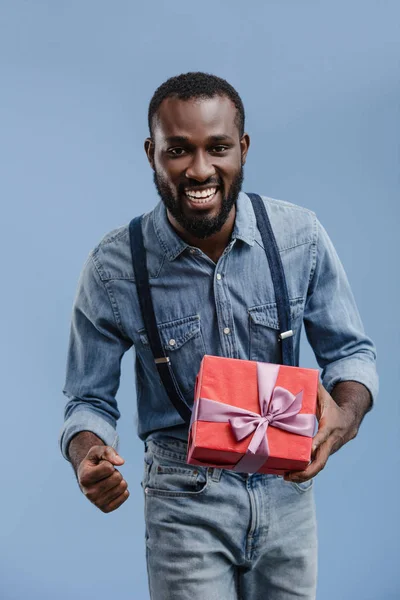 Excité jeune homme afro-américain avec boîte cadeau enveloppé par un ruban geste à la main isolé sur fond bleu — Photo de stock