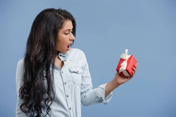 Side view of shocked african american girl looking at red gift box isolated on blue — Stock Photo