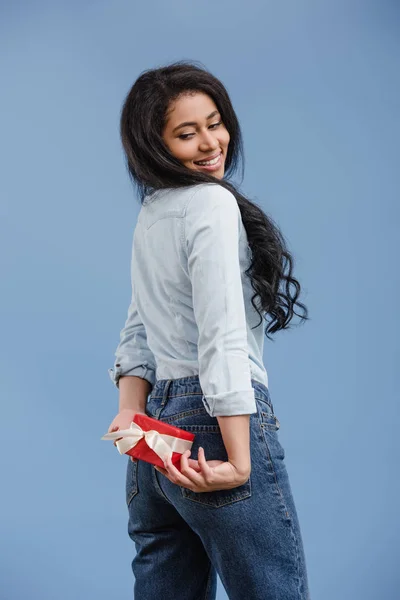 Sonriente atractivo afroamericano chica sosteniendo rojo regalo caja aislado en azul - foto de stock