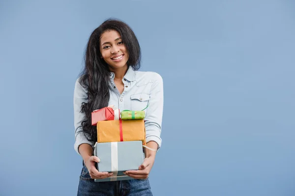 Sonrisa atractiva afroamericana chica sosteniendo cajas de regalo aislado en azul - foto de stock