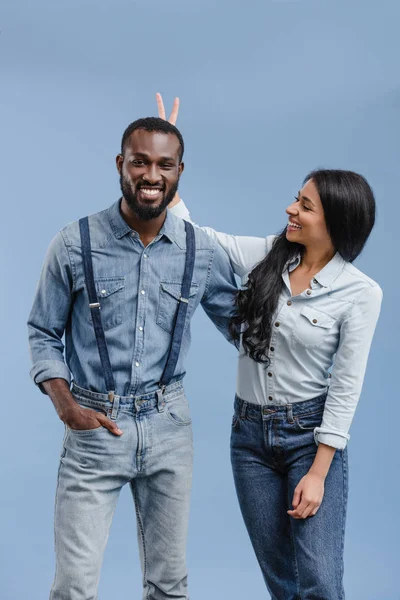 Smiling african american girlfriend showing two fingers above boyfriend head isolated on blue — Stock Photo