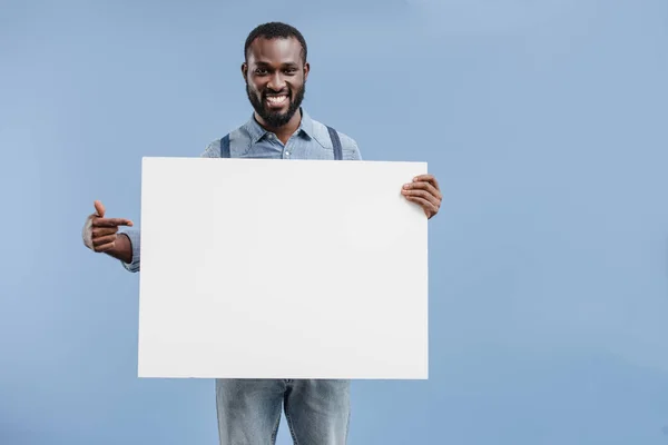 Sonriente guapo afroamericano hombre apuntando en blanco cartel aislado en azul - foto de stock