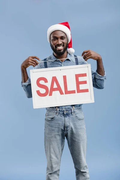 Smiling handsome african american man in santa hat holding sale sign isolated on blue — Stock Photo