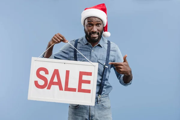 Grimacing handsome african american man in santa hat pointing on sale sign isolated on blue — Stock Photo