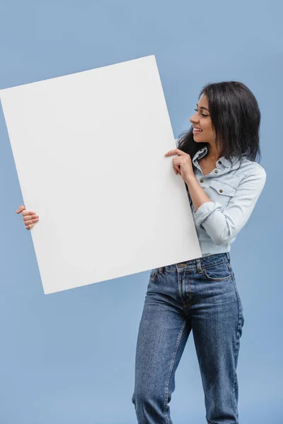Sorrindo atraente menina afro-americana olhando para cartaz em branco isolado em azul — Fotografia de Stock