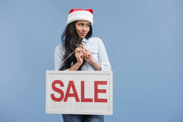 Cheerful attractive african american girl in santa hat holding sale sign isolated on blue — Stock Photo