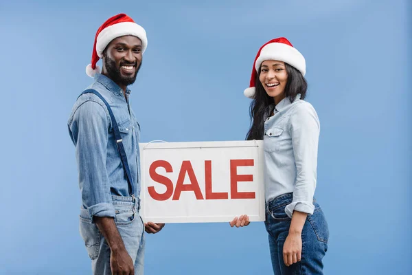 Smiling african american couple in santa hats holding sale sign isolated on blue — Stock Photo