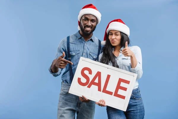 Afrikanisch-amerikanisches Paar in Weihnachtsmannmützen zeigt auf Verkaufsschild isoliert auf blau — Stockfoto