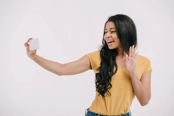 Atractiva chica afroamericana agitando la mano durante la videollamada en el teléfono inteligente aislado en blanco - foto de stock