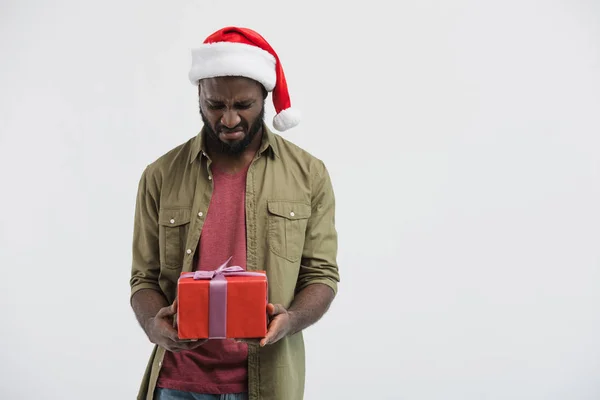 Sconvolto afroamericano uomo in cappello di Babbo Natale guardando scatola presente isolato su bianco — Foto stock