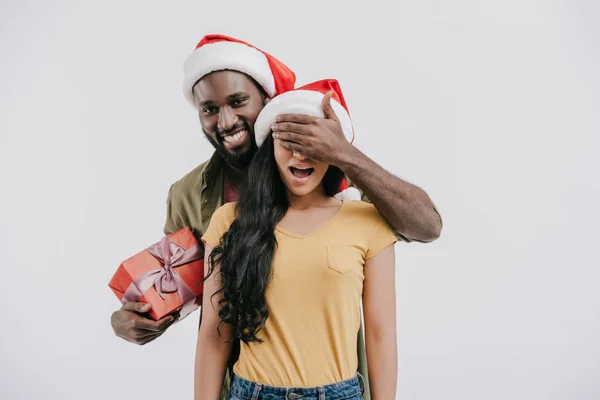 Афроамериканський хлопець закриває очі дівчині і тримає подарунок на Новий рік ізольовано на білому — стокове фото