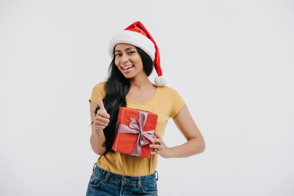 Lächelndes afrikanisch-amerikanisches Mädchen mit Weihnachtsmütze, Geschenk in der Hand und Daumen hoch auf weiß — Stockfoto