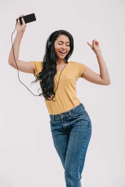 Atractiva chica afroamericana bailando mientras escucha música con teléfono inteligente y auriculares aislados en blanco - foto de stock
