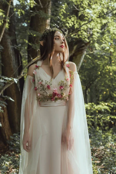 Attraktive mystische Elfe in Blumenkranz und Kleid mit Blumen im Wald — Stockfoto