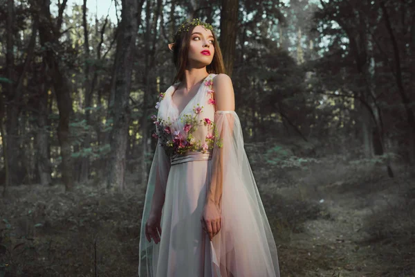 Привлекательная мистическая девушка с эльфийскими ушами, позирующая в цветочном платье в лесу — стоковое фото