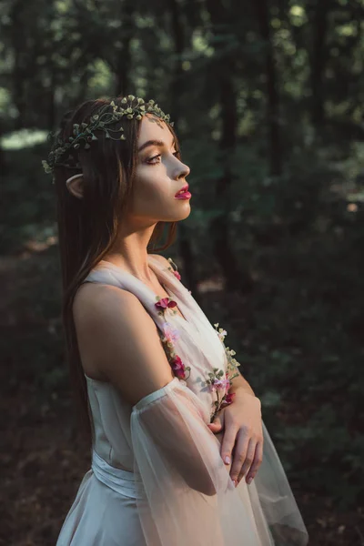 Elfo místico atraente em vestido de flor e grinalda de pé com braços cruzados na floresta — Fotografia de Stock