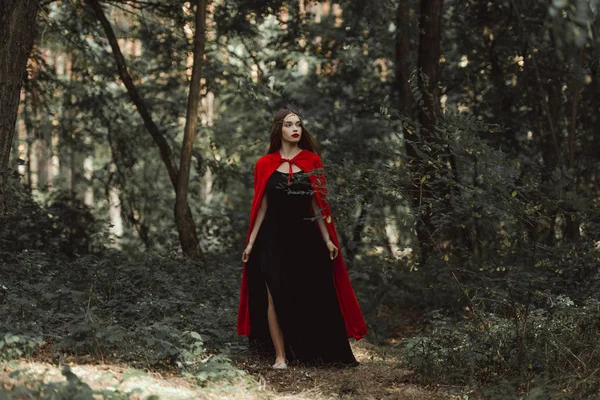 Chica mística en vestido negro y capa roja caminando en el bosque - foto de stock