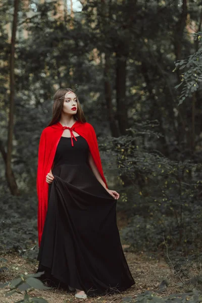 Attractive mystic woman in black dress and red cloak walking in woods — Stock Photo