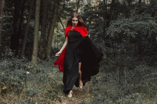 Elegante ragazza mistica in abito nero e mantello rosso in esecuzione nei boschi — Foto stock